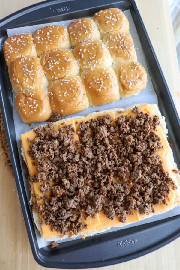 A baking tray with pull-apart slider buns. The top half shows buns with sesame seeds; the bottom half has open buns layered with sliced cheese and cooked ground beef.