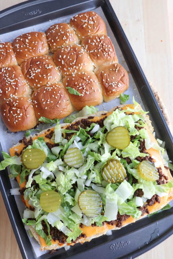 A baking tray with pull-apart sesame seed slider buns; half the tray shows the top of the buns, the other half shows open buns topped with melted cheese, ground beef, lettuce, onions, and sliced pickles.