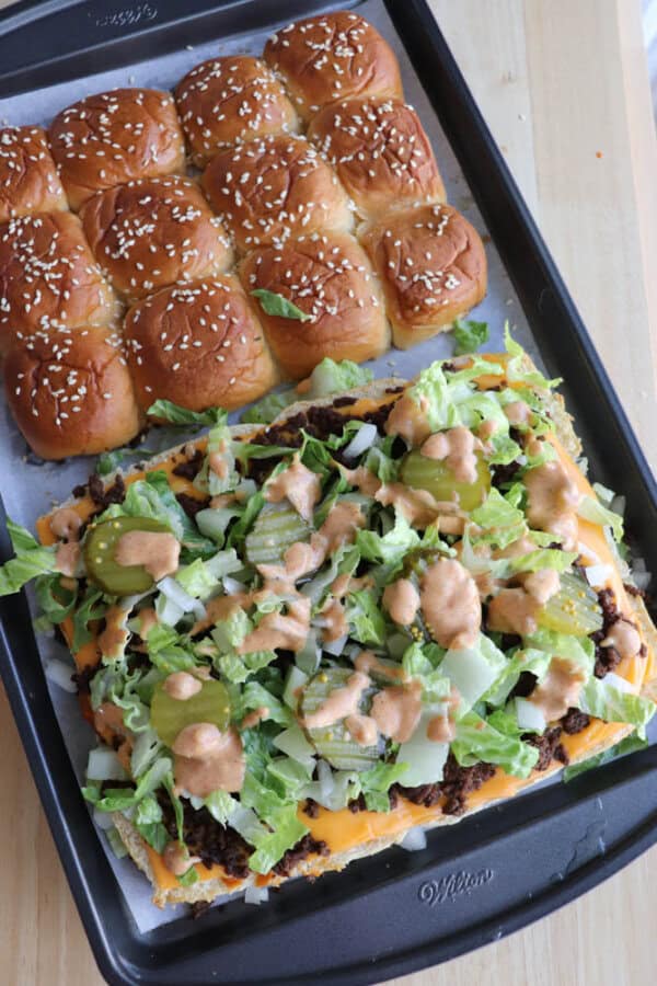 A baking tray with two sections: one with golden, sesame seed-topped slider buns, and the other with sliders topped with ground beef, melted cheese, lettuce, pickles, onions, and sauce.
