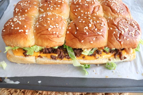 A tray of pull-apart cheeseburger sliders made with sesame seed buns, ground beef, cheddar cheese slices, and shredded lettuce, placed on parchment paper.