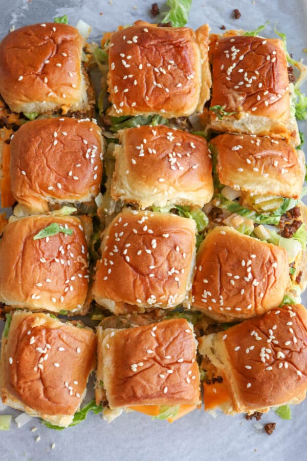 Twelve mini cheeseburgers with sesame seed buns are arranged closely together on a tray, topped with lettuce, cheese, and visible ground beef patties.