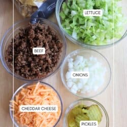 Six bowls containing nachos, shredded lettuce, cooked ground beef, chopped onions, shredded cheddar cheese, and sliced pickles are arranged on a table, viewed from above.