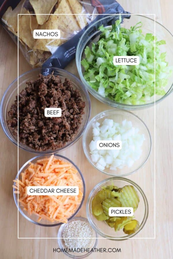 Six bowls containing nachos, shredded lettuce, cooked ground beef, chopped onions, shredded cheddar cheese, and sliced pickles are arranged on a table, viewed from above.