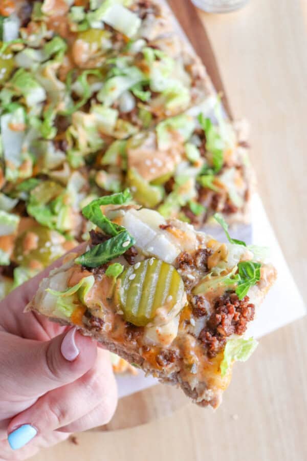 A hand holding a slice of pizza topped with ground meat, lettuce, onions, pickles, sesame seeds, and a drizzle of sauce, with more pizza in the background on a wooden surface.