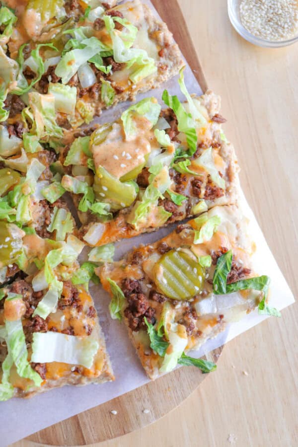A close-up of sliced pizza topped with ground beef, melted cheese, lettuce, chopped onions, pickles, and a drizzle of sauce, placed on parchment paper on a wooden surface. A small dish of sesame seeds is nearby.
