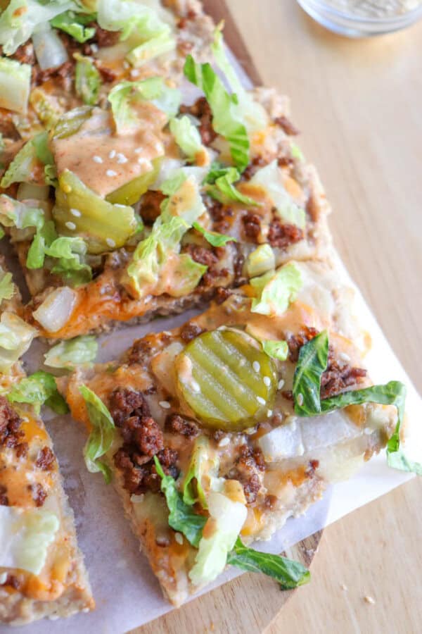 A close-up of pizza slices topped with ground meat, shredded lettuce, pickles, onions, sesame seeds, and a creamy orange sauce on a wooden surface.