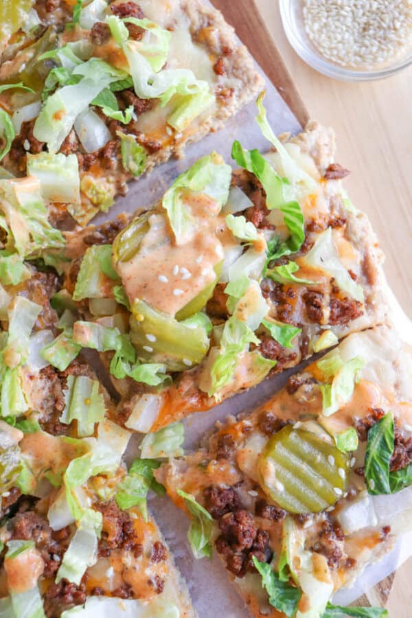 A close-up of pizza slices topped with ground beef, shredded lettuce, pickles, diced onions, and an orange sauce, with a small bowl of sesame seeds in the background.