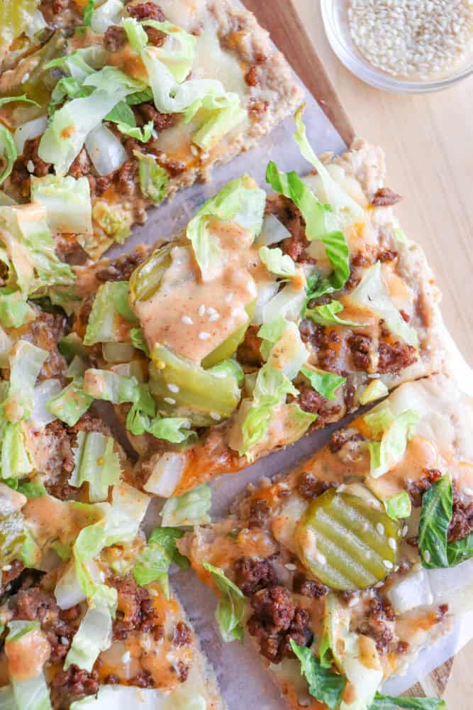 A close-up of pizza slices topped with ground beef, shredded lettuce, pickles, diced onions, and an orange sauce, with a small bowl of sesame seeds in the background.