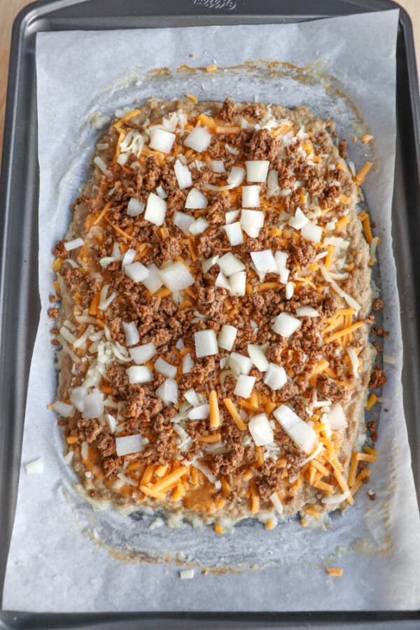 Unbaked pizza on parchment paper-lined baking sheet, topped with shredded cheese, diced onions, and crumbled cooked ground meat, ready to go in the oven.