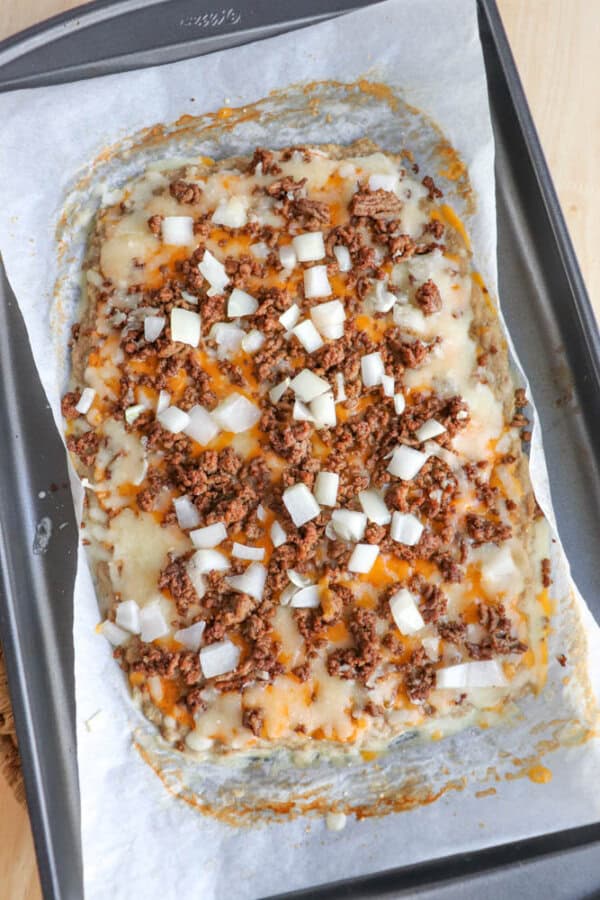 A rectangular sheet of cheesy, baked food topped with ground beef and chopped raw onions sits on parchment paper on a baking tray. The edges are slightly browned and melted cheese is visible.