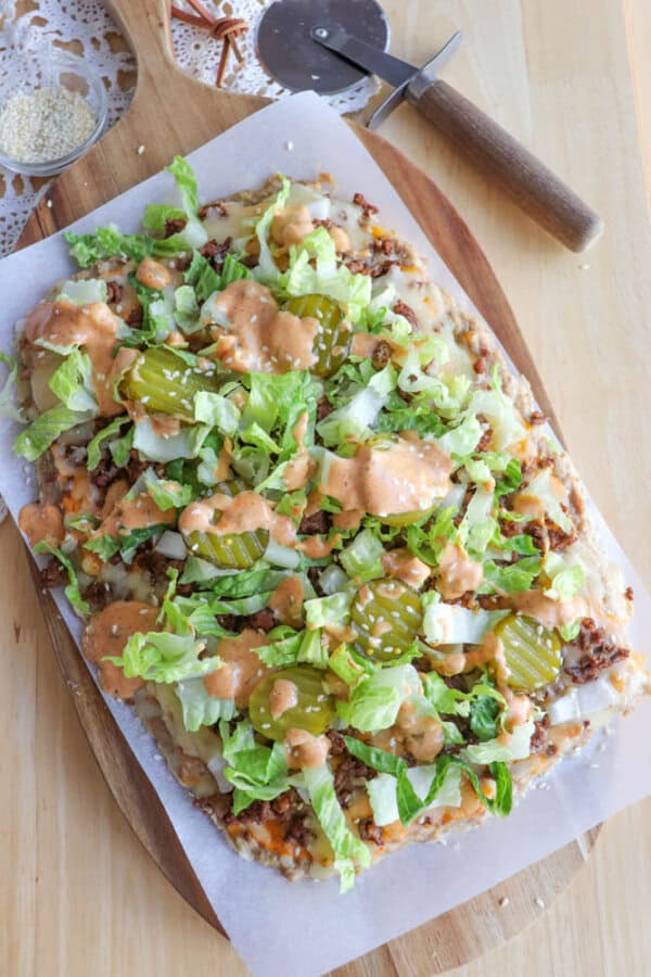 A rectangular pizza topped with shredded lettuce, pickles, diced onions, ground meat, and a creamy sauce on parchment paper, placed on a wooden board with a pizza cutter beside it.