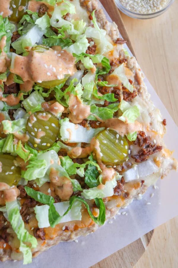 A close-up of a pizza topped with shredded lettuce, sliced pickles, diced onions, sesame seeds, melted cheese, and a drizzle of creamy sauce on a wooden surface.