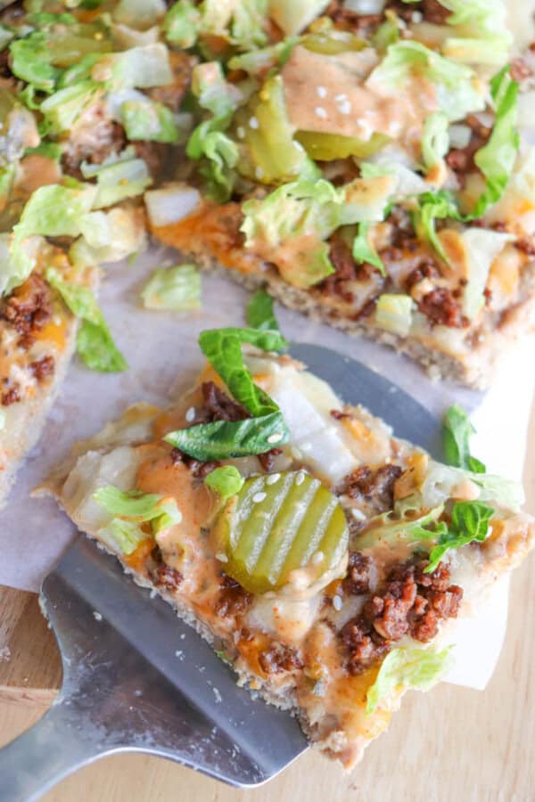 A close-up of a square slice of cheeseburger pizza on a spatula, topped with ground beef, lettuce, pickles, cheese, onions, and sauce. The rest of the pizza is visible in the background.