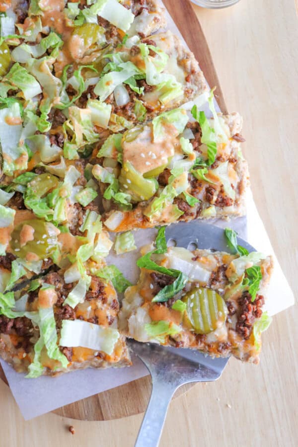 A rectangular pizza topped with ground beef, melted cheese, lettuce, pickles, diced onions, and creamy sauce. A slice is being lifted from the pizza with a metal spatula.