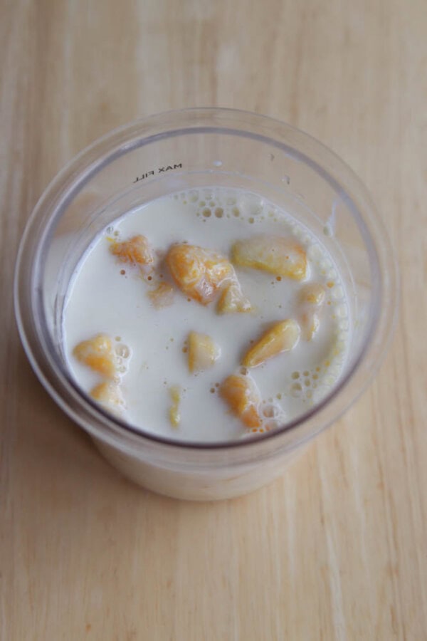 A clear cup filled with milk and chunks of yellow fruit, possibly banana, sits on a light wooden surface.