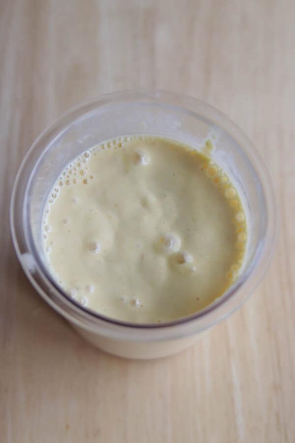 A close-up view of a glass filled with a thick, pale yellow smoothie or milkshake, with small bubbles on the surface, placed on a light wooden surface.