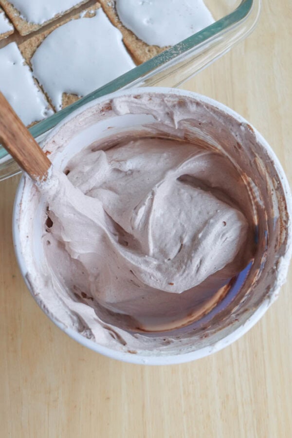 A bowl of light, fluffy chocolate mousse being mixed with a wooden spoon, with a glass dish of frosted graham cracker squares visible in the background.