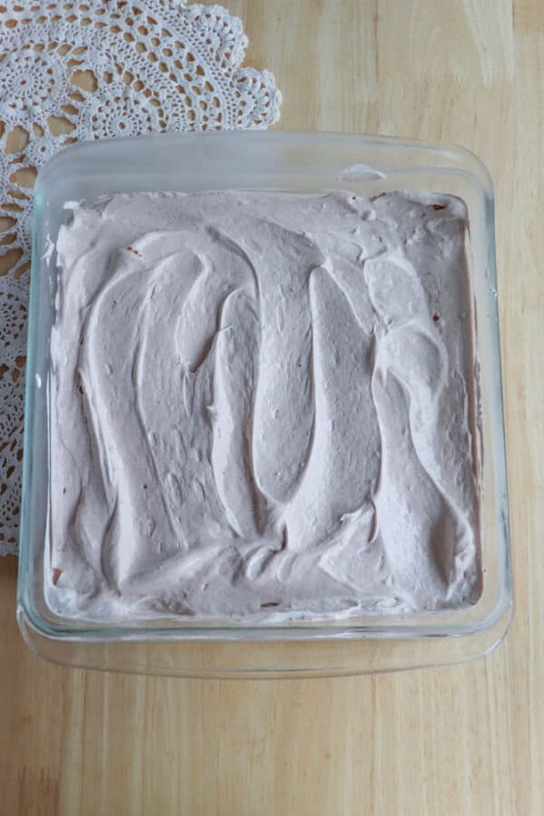 A glass baking dish filled with a dessert topped with swirled, light brown whipped topping sits on a wooden surface next to a white lace doily.