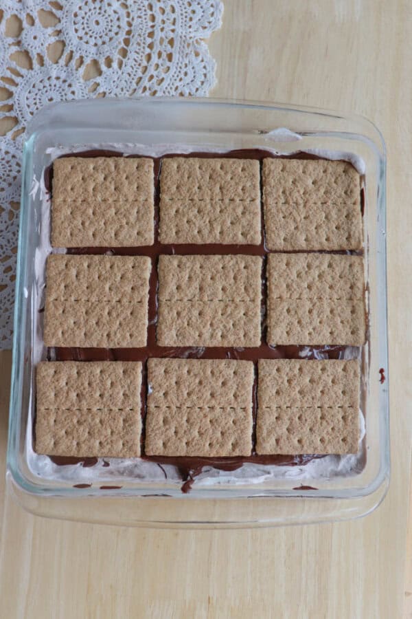 A glass baking dish filled with a layered dessert made of graham crackers, chocolate, and marshmallow, resembling s’mores. The top layer shows nine neatly arranged rectangular graham crackers.