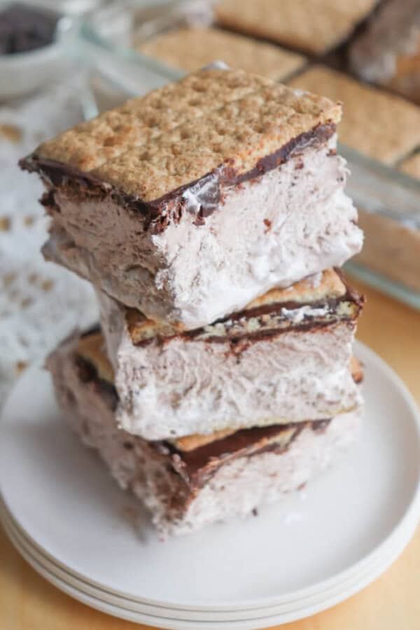Three ice cream sandwiches made with graham crackers, chocolate, and marshmallow ice cream are stacked on a white plate, with more sandwiches visible in the background.