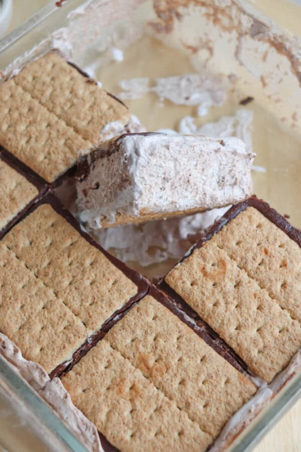 A close-up of a glass dish filled with ice cream sandwiches made from graham crackers, chocolate, and a creamy filling. One sandwich is being lifted out, showing its layers.