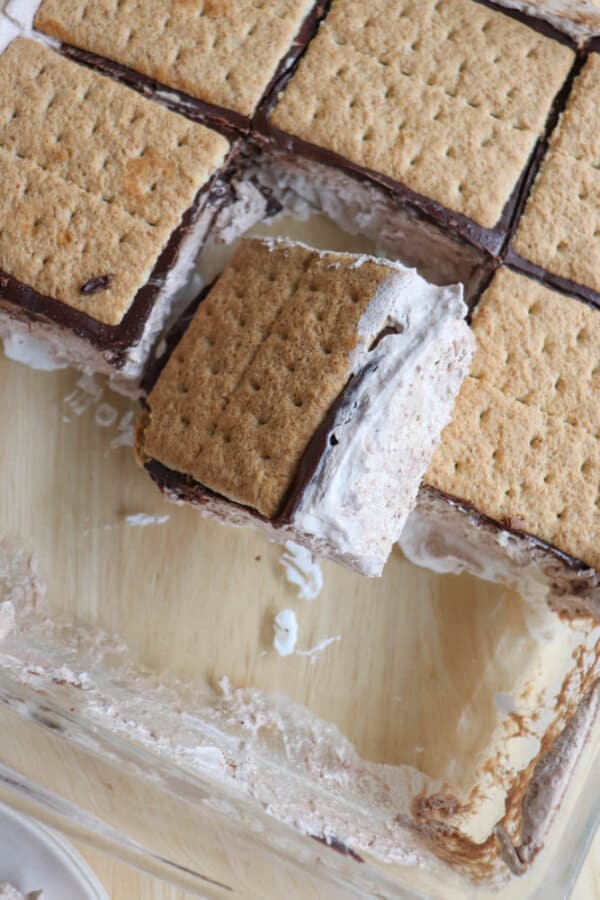 A close-up of homemade ice cream sandwiches in a glass dish, made with graham crackers, chocolate, and a creamy filling. One sandwich is lifted out, showing the layers and creamy texture inside.