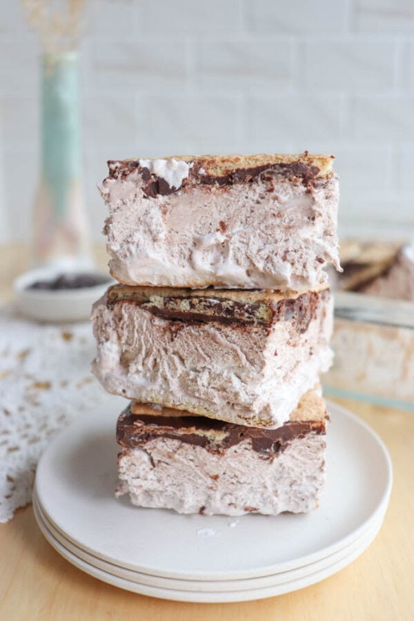 Three large smores ice cream sandwiches stacked on white plates, filled with creamy ice cream and chocolate layers between graham crackers, with a blurred dish and vase in the background.