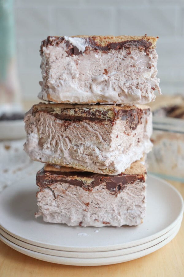 Three thick ice cream sandwiches with chocolate and graham cracker layers are stacked on a white plate, with creamy filling and slightly messy edges.