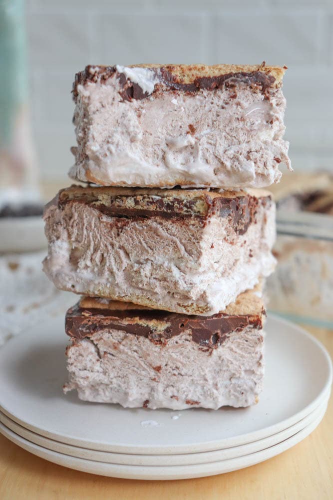 Three thick ice cream sandwiches with chocolate and graham cracker layers are stacked on a white plate, with creamy filling and slightly messy edges.
