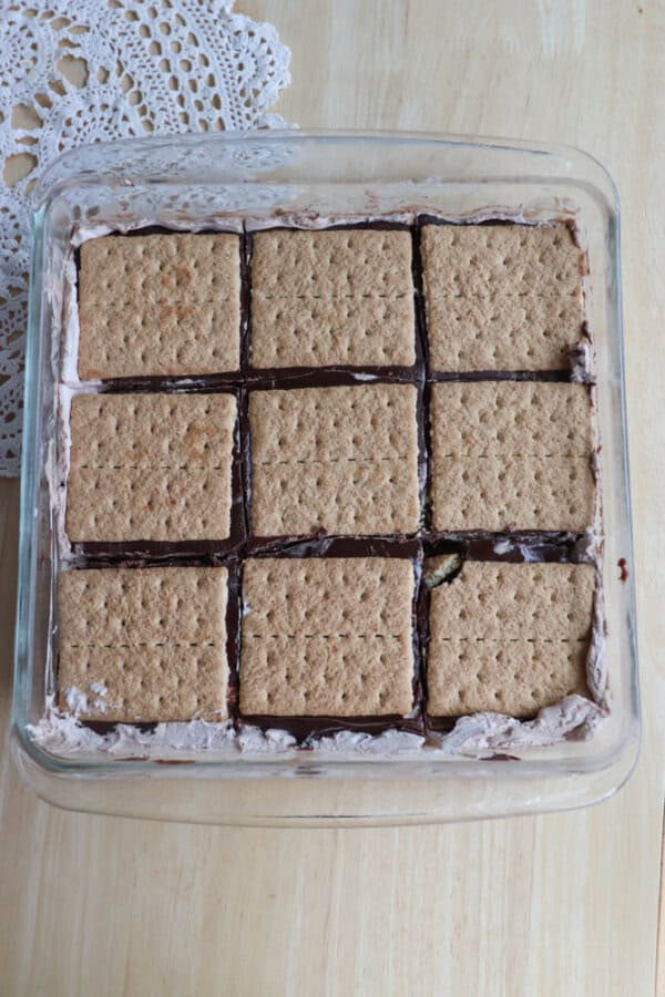 A glass baking dish filled with nine neatly arranged squares of dessert, each topped with a graham cracker. Chocolate and cream layers are visible under the crackers. A crochet doily is partially visible on the side.