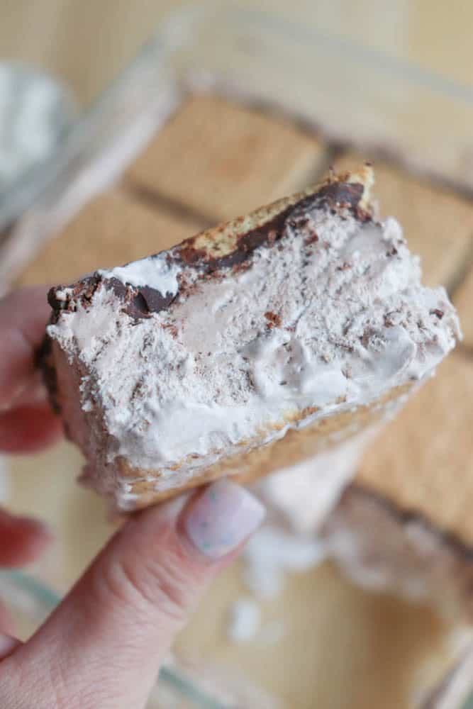 A hand holding a partially eaten ice cream sandwich made with graham crackers, a chocolate layer, and creamy filling. More sandwiches are visible in the background.
