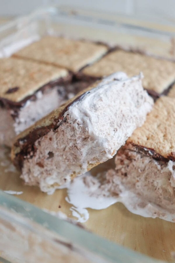 A close-up of homemade ice cream sandwiches in a glass dish, featuring thick layers of creamy filling between crumbly graham cracker cookies, with some melting and gooey texture visible.
