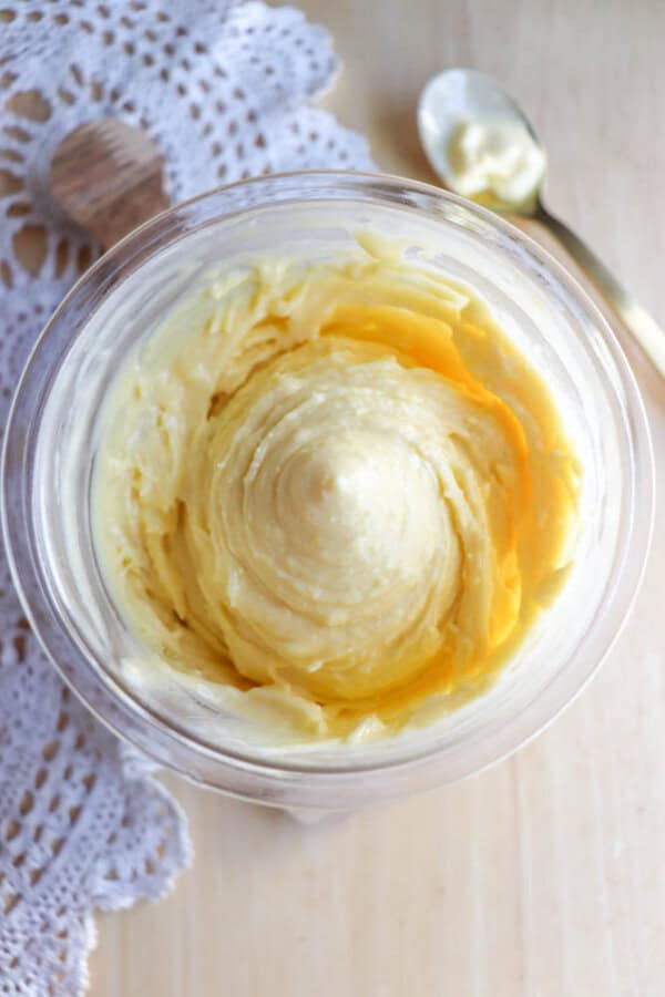 A close-up top view of creamy, yellow homemade butter in a glass jar, with a gold spoon and a white lace cloth nearby on a light wooden surface.