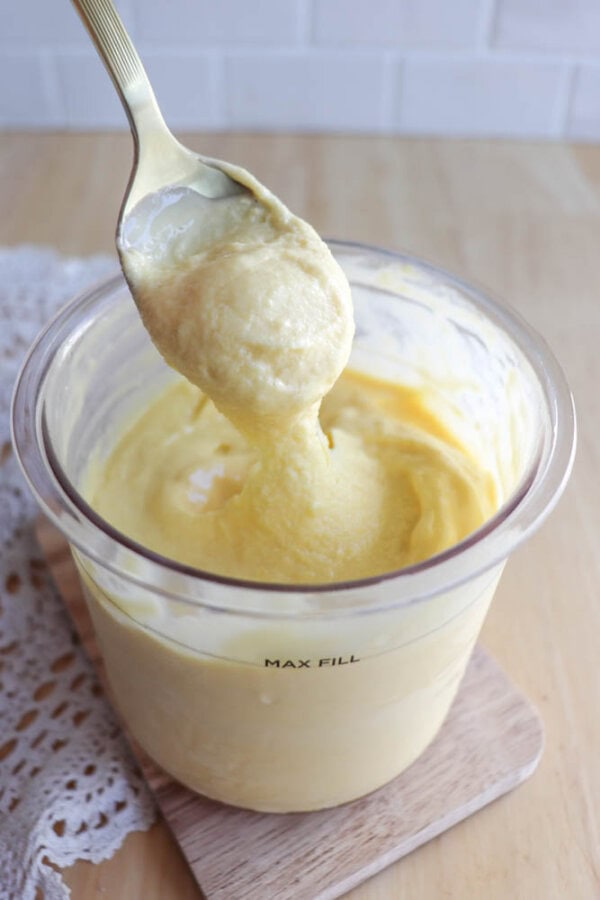 A spoon holding thick, creamy yellow batter or custard above a clear container filled with the same mixture, placed on a wooden board with a lace cloth nearby.