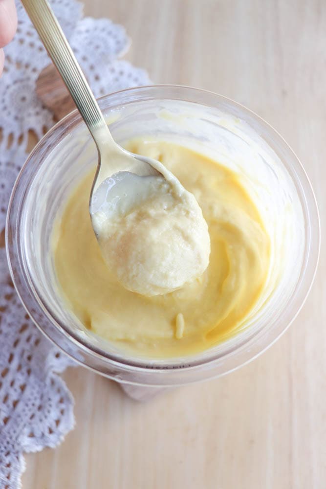 A spoon scoops thick, creamy yellow custard from a clear plastic cup, placed on a light wooden surface next to a white lace cloth.