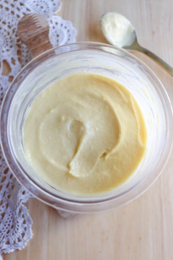 A glass bowl filled with creamy yellow custard sits on a wooden surface next to a metal spoon and a piece of white lace fabric.