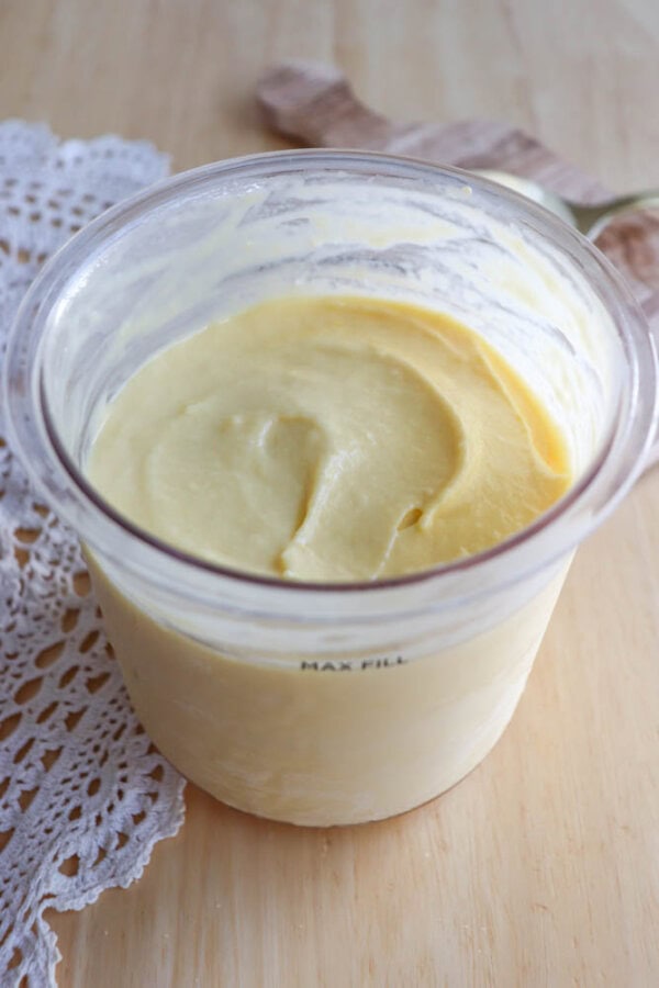 A glass container filled with creamy, yellow homemade butter sits on a wooden surface next to a white lace cloth.