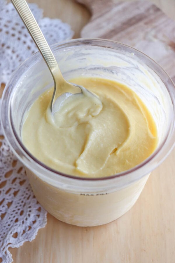A clear container filled with creamy, pale yellow custard or pudding sits on a wooden surface next to a lace cloth. A spoon rests inside, showing the smooth, thick texture of the mixture.