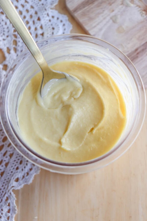A glass bowl filled with creamy yellow custard or pudding, with a silver spoon resting inside. The bowl is on a wooden surface next to a white lace cloth.