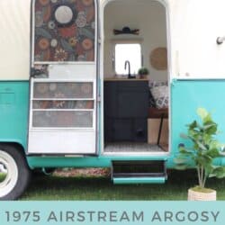 A vintage turquoise and white 1975 Argosy Airstream camper with an open door, floral screen, striped awning, and potted plant outside. Peek inside this cozy argosy airstream restoration featuring black cabinetry and decor.