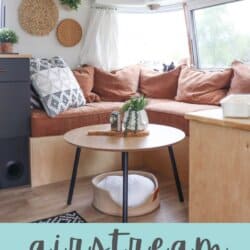 Cozy renovated Argosy Airstream interior with a brown sectional sofa, geometric pillows, round wooden coffee table, and woven wall decor. Large windows fill this Airstream restoration with natural light. Text: โAirstream 1975 Restoration homemadewithheather.comโ.