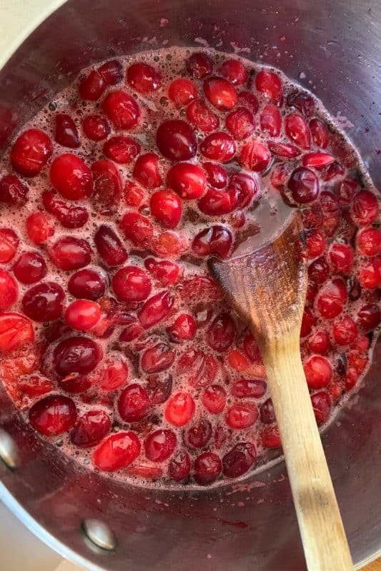 A pot of cranberries bubbling in liquid, being stirred with a wooden spoon, likely as they cook down to make cranberry sauce.