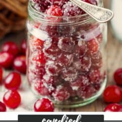 A glass jar filled with sugar-coated candied cranberries, with a small silver spoon resting inside. Fresh cranberries are scattered around the jar on a wooden surface. Text reads: “candied cranberries homemadeheather.com”.