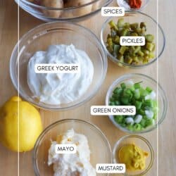 Bowls of ingredients for potato salad are arranged on a wooden surface, including baby potatoes, Greek yogurt, mayo, mustard, oil, chopped pickles, green onions, spices, and a whole lemon.