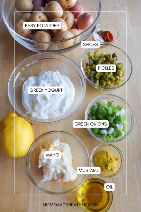 Bowls of ingredients for potato salad are arranged on a wooden surface, including baby potatoes, Greek yogurt, mayo, mustard, oil, chopped pickles, green onions, spices, and a whole lemon.