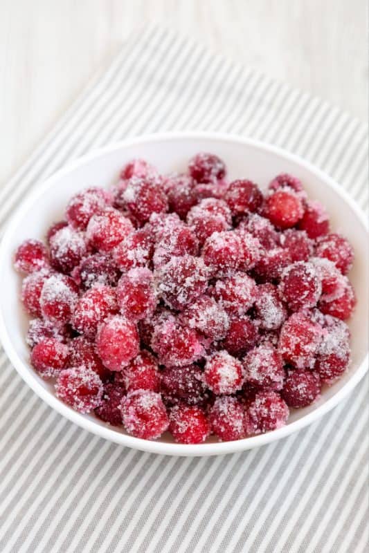 A white bowl filled with cranberries coated in sugar, placed on a white and gray striped cloth.