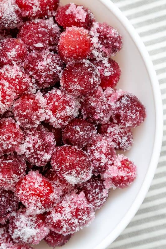 Sugared raspberries in a white bowl, coated with a layer of granulated sugar, placed on a surface with gray and white stripes.