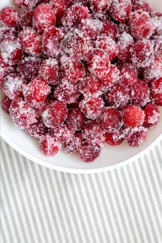 A white bowl filled with bright red cranberries coated in sugar, placed on a white and gray striped surface.