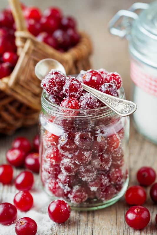 A glass jar filled with sugared cranberries, with a vintage silver spoon resting on top. Fresh cranberries and a wicker basket are in the background on a rustic wooden surface.