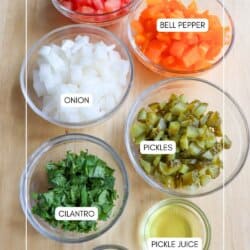 Seven glass bowls on a wooden surface contain labeled ingredients: chopped tomato, bell pepper, onion, pickles, cilantro, dill, and a small amount of pickle juice.